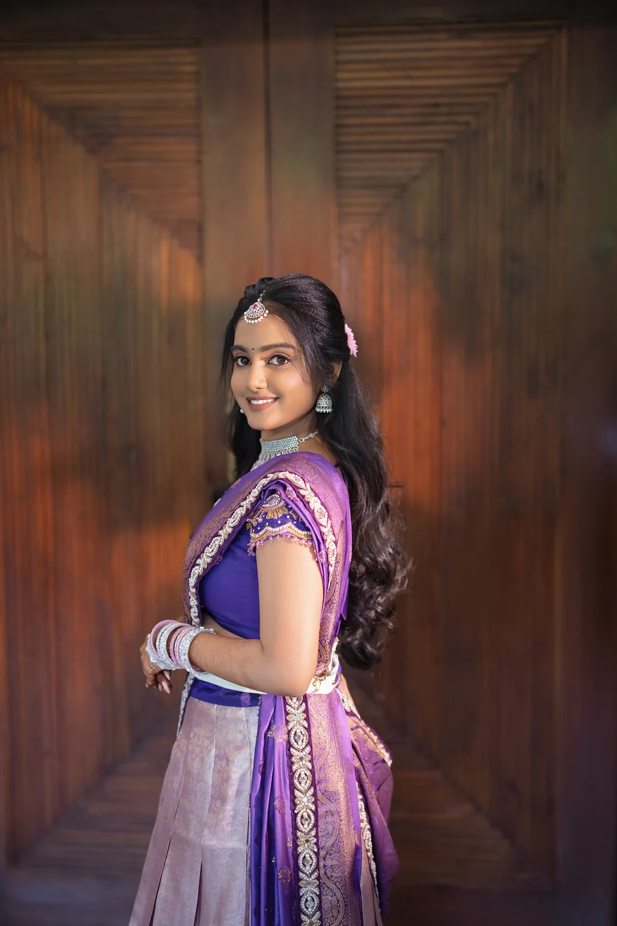 Harini Veena smiling in a lavender and purple half-saree against a wooden wall