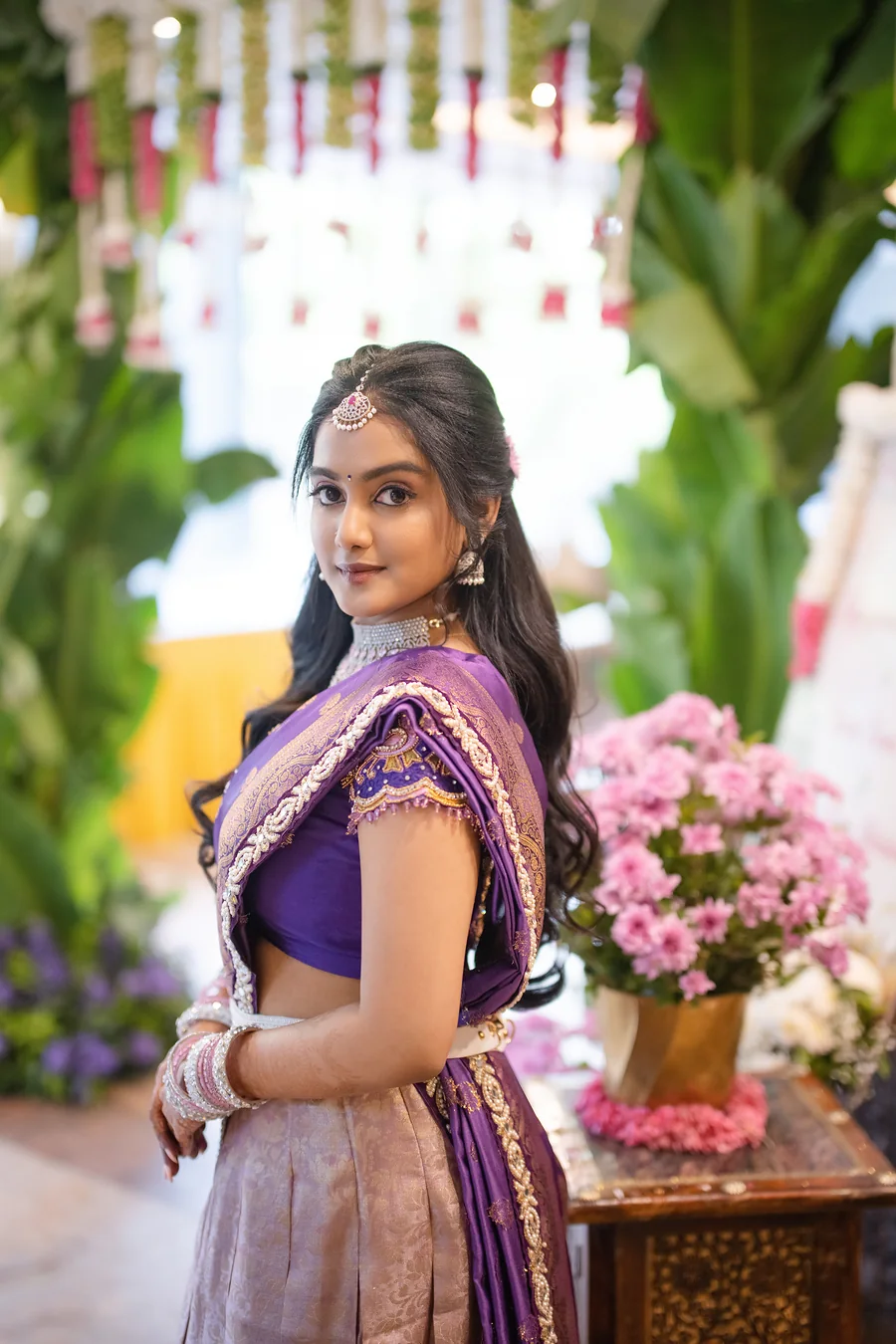Harini Veena in a lavender and purple half-saree with a maang tikka, among pink flowers