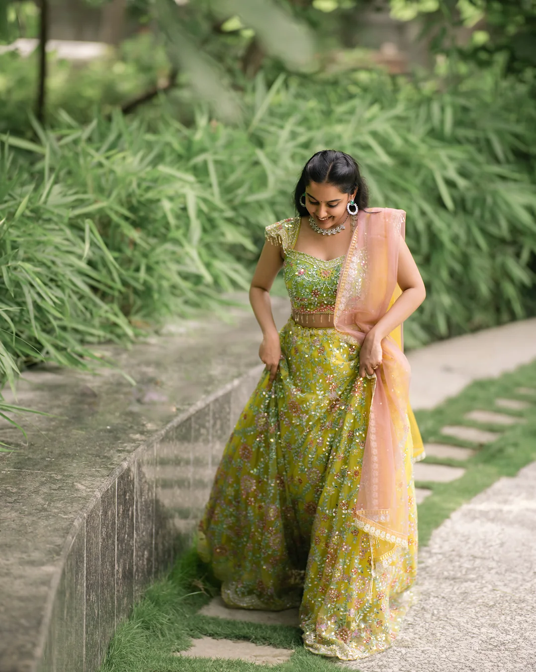 Harini Veena walking along a garden path in a lime lehenga with a peach dupatta