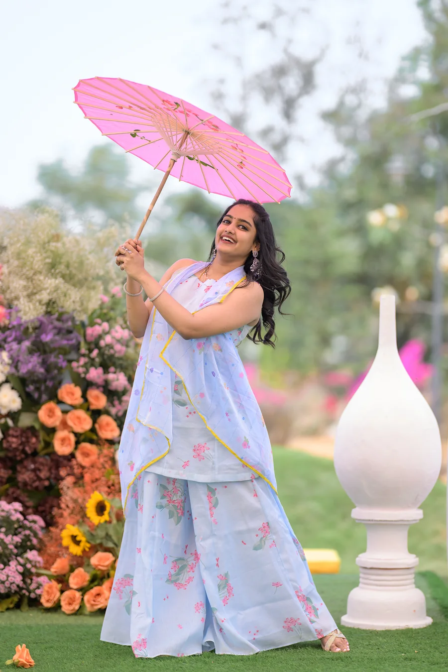 Harini Veena smiling under a pink paper parasol in a pastel blue floral outfit